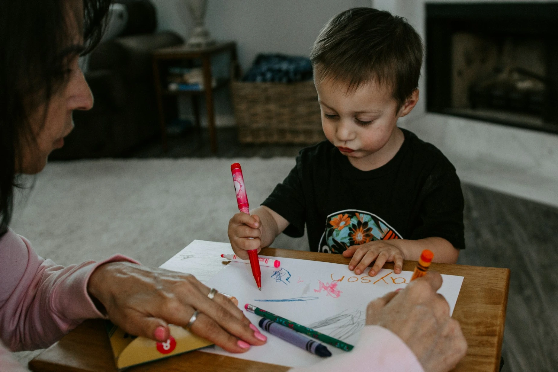 boy in black neck t-shirt holding pink pen writing on white paper